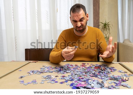 Handsome bearded man in an orange sweater doing puzzle with a worried and concentrated gesture. Entertainment concept