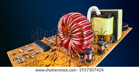 Electronic components in orange circuit board flying on dark blue background in abstract scene. Ferrite core coils, resistors and a chip or transformer on PCB detail in panoramic artistic still life. Royalty-Free Stock Photo #1937307424