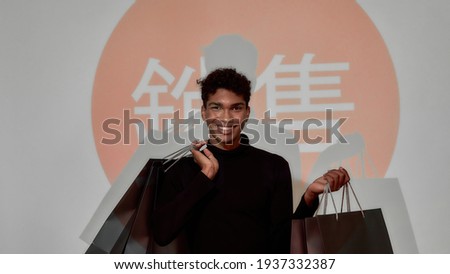 Cheerful young african american man with bags looking at camera while standing in front of screen with japanese flag and hieroglyphs, widescreen. Discount and sale concept