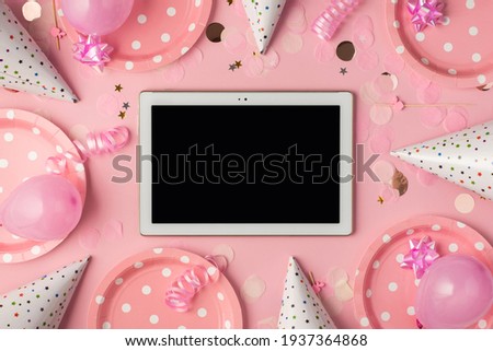 Top view photo of birthday composition with pink ribbon stars serpentine hats paper plates balloons confetti and tablet computer in the middle with copyspace on pastel pink background