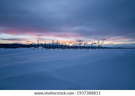 Winter frozen lake scene in northern Canada on a stunning cloudy sunset afternoon in March with white snow, mountains in background and iconic Canadian landscape in the north.  Royalty-Free Stock Photo #1937388811