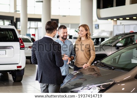 Young car salesman showing to mid-age couple new automobile at dealership salon. Royalty-Free Stock Photo #1937414719