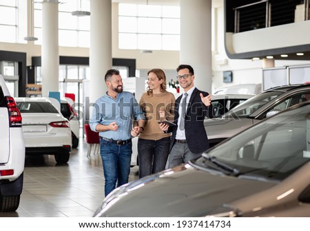 Young car salesman showing to mid-age couple new automobile at dealership salon. Royalty-Free Stock Photo #1937414734