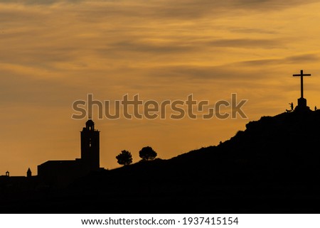 silhouette of people and dog on the top of a hill with a large cross with a church and trees in the background during the last lights of dusk