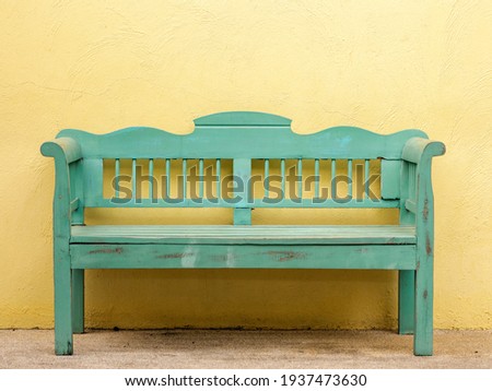 The old wooden bench in green color in front of the yellow wall. Wooden 2 seater garden bench. The green wooden bench and the yellow wall. Garden or outdoor furniture. Royalty-Free Stock Photo #1937473630