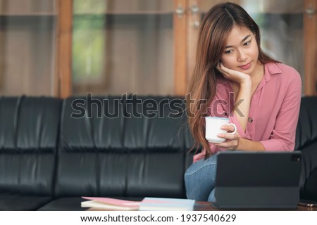Pretty asian woman holding cup of coffee and looking at tablet screen while sitting on sofa in living room. Work from home concept.