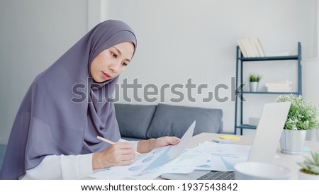 Beautiful Asia muslim lady in headscarf casual wear using laptop in living room at house. Remotely working from home, new normal lifestyle, social distancing, quarantine for corona virus prevention.