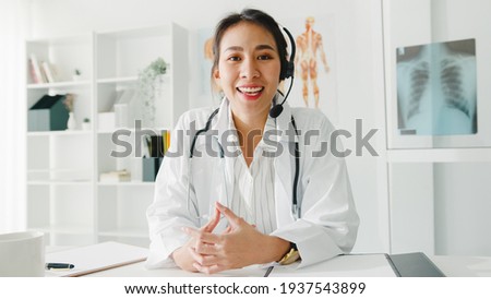 Young Asia lady doctor in white medical uniform with stethoscope using computer laptop talk video conference call with patient, looking at camera in health hospital. Consulting and therapy concept. Royalty-Free Stock Photo #1937543899