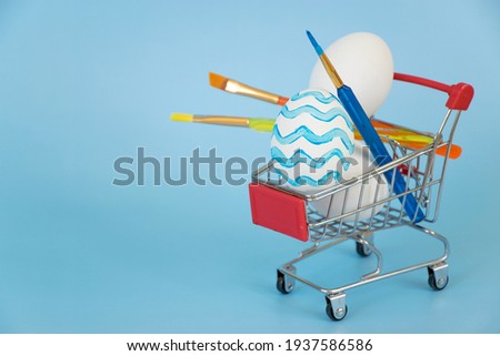 Easter egg decorated in blue waves with other white eggs and paintbrushes in shopping cart on blue background. Preparation and shopping for Happy Easter holiday with copy space.