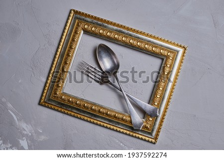 Old, iron, fork and spoon in a gilded frame against a gray concrete wall.