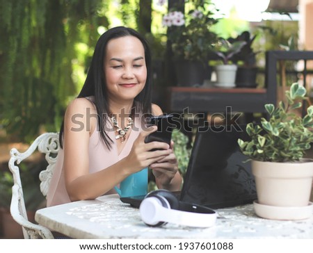 Portrait of Asian woman  sitting with blue coffee cup and computer notebook on table in her home garden, using mobile phone,  smiling and looking at mobile phone screen.