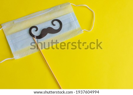 Medical face mask, paper black mustache on a stick on bright yellow background, close-up. Celebrating April Fools ' Day during the coronavirus pandemic.