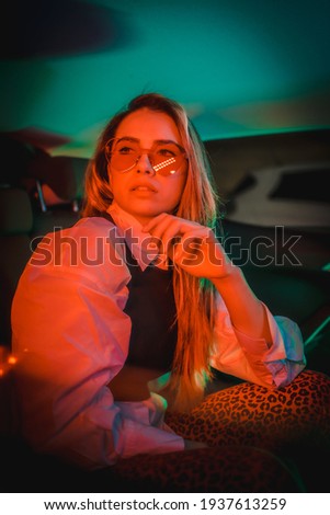 Urban photography with red and green neons in the back of a car of a young blond Caucasian woman with heart glasses