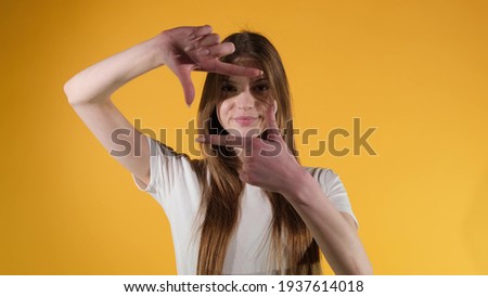 girl frames her face with her fingers, pretending to take a picture, orange studio background