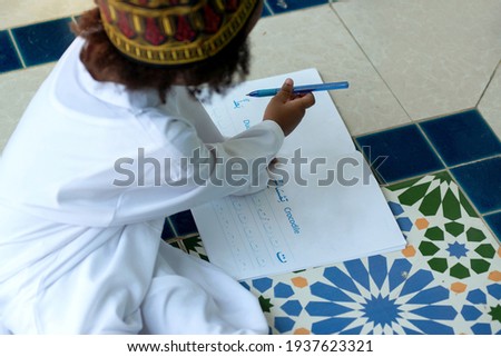 Child boy wearing a traditional Muslim suit  learning the Arabic alphabet, Children write the names of the animals for practice in the book (Arabic text mean crocodile and duck ), View from above
