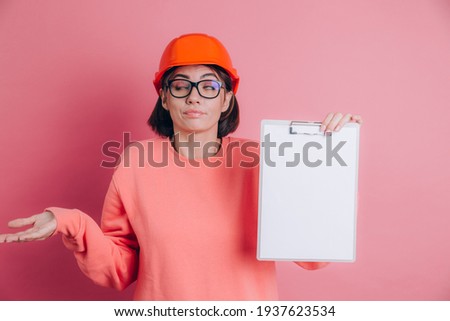 Woman worker builder hold white sign board blank against pink background. Building helmet. Confused seeks a solution. Female throwing up her hands and shrugging shoulders, having no clue