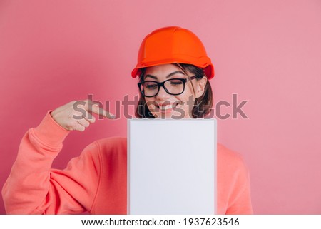 Smiling woman worker builder hold white sign board blank against pink background. Building helmet. Point index finger.