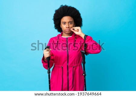 Young Africa American with backpack and trekking poles isolated on blue background showing a sign of silence gesture
