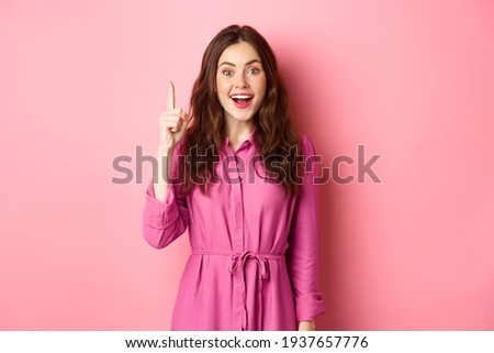 Excited beautiful girl with make up on, having an idea, raising finger up in eureka sign and smiling, saying her plan, standing against pink background