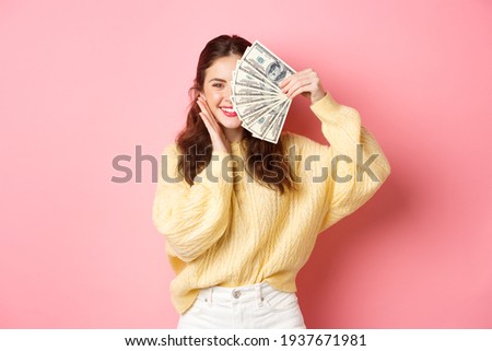 Shopping. Beautiful brunette woman holding money on face and smiling, touching cheek, standing against pink background Royalty-Free Stock Photo #1937671981