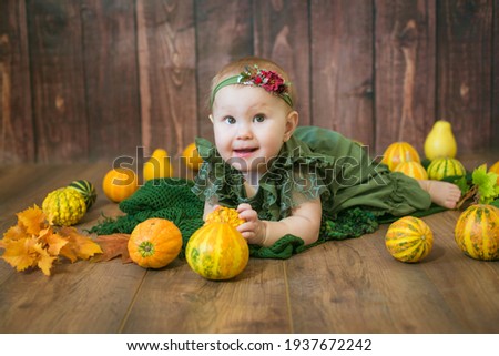 Cute little girl up to 1 year old in a green cute dress and a floral wreath with small yellow and orange pumpkins on a brown wooden background 