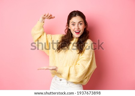Young happy woman staring at camera, showing big object, holding your logo or product with her hands aside on copy space, standing against pink background Royalty-Free Stock Photo #1937673898