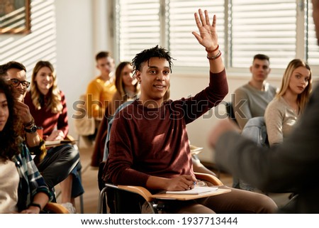 Smiling African American student raising his hand to ask a questing during a class at lecture hall.  Royalty-Free Stock Photo #1937713444