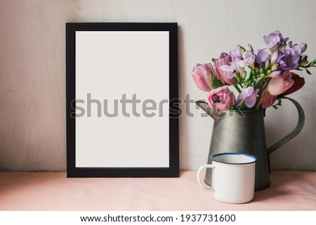 Black frame mockup with a bouquet of spring flowers in vintage vase and coffee mug on a table. Portrait orientation. Retro styled desk with copy space template. 