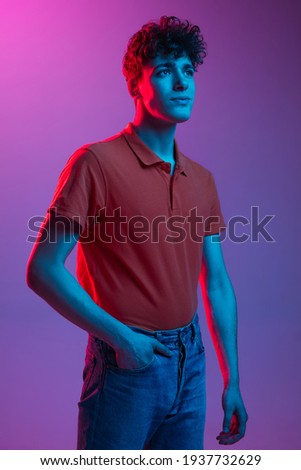 Young Caucasian man in bright clothes red shirt and jeans posing isolated on multicolored background in neon light. Concept of human emotions, facial expression, youth culture. Copy space for ad.