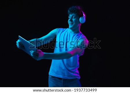 Young Caucasian man in headphones using digital tablet isolated on dark background in neon light. Human emotion, facial expressions, youth culture, moder life. Copy space for ad, design.