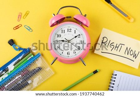 A table clock with Right now words Written on a sticky note with other elements on a Yellow background flat lay shot