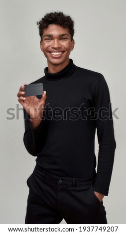 Happy young african american man holding plastic card mockup and smiling at camera while posing on light background, vertical shot. Discount and sale concept