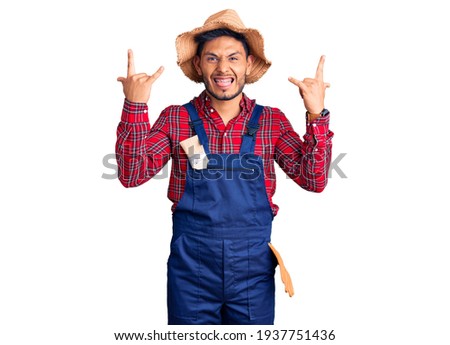 Handsome latin american young man weaing handyman uniform shouting with crazy expression doing rock symbol with hands up. music star. heavy music concept. 