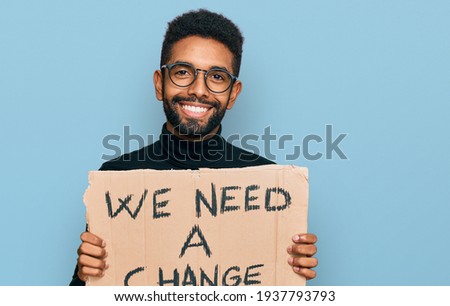 Young african american man holding we need a change banner looking positive and happy standing and smiling with a confident smile showing teeth 