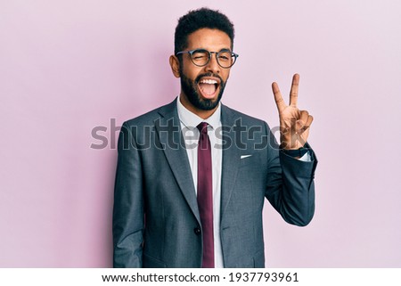 Handsome hispanic business man with beard wearing business suit and tie smiling with happy face winking at the camera doing victory sign. number two. 