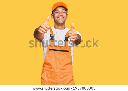 Young handsome african american man wearing handyman uniform approving doing positive gesture with hand, thumbs up smiling and happy for success. winner gesture. 