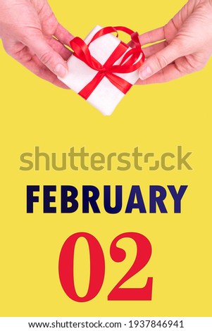 February 2nd. Festive Vertical Calendar With Hands Holding White Gift Box With Red Ribbon And Calendar Date 2 February On Illuminating Yellow Background. Winter month, day of the year concept.