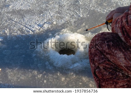 A fisherman in warm winter clothes catches fish on a spinning rod in a hole in the ice of the river in winter.
