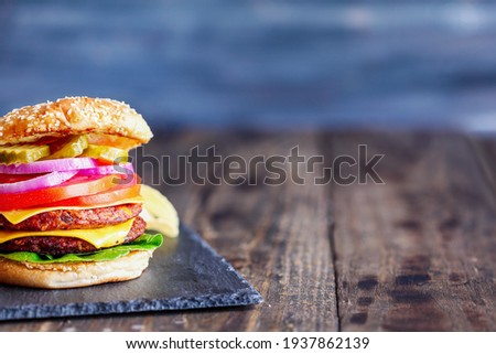 Vegetarian cheeseburger made with two meat substitute patties, slices of melted cheese, onions, pickles, lettuce, and tomato on a fresh sesame seed bun over a rustic dark background. 
