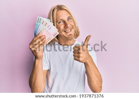 Caucasian young man with long hair holding new taiwan dollars banknotes smiling happy and positive, thumb up doing excellent and approval sign 