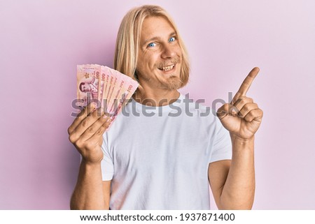 Caucasian young man with long hair holding 100 thai baht banknotes smiling happy pointing with hand and finger to the side 