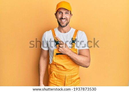 Young handsome man wearing handyman uniform over yellow background cheerful with a smile on face pointing with hand and finger up to the side with happy and natural expression 