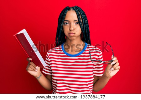 Beautiful hispanic woman reading a book with glasses puffing cheeks with funny face. mouth inflated with air, catching air. 
