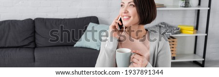 Cheerful woman with cup talking on smartphone in living room, banner