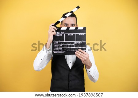 Young man with beard wearing bow tie and vest holding clapperboard very happy having fun