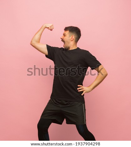 Muscle building. Young Caucasian man in black sports attire standing isolated over pink background. Concept of human emotions, facial expression, funny meme emotions, sports. Copyspace for ad.