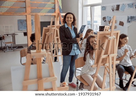 Portrait of smiling female arfican american teacher in modern classroom with kids drawing lesson in background