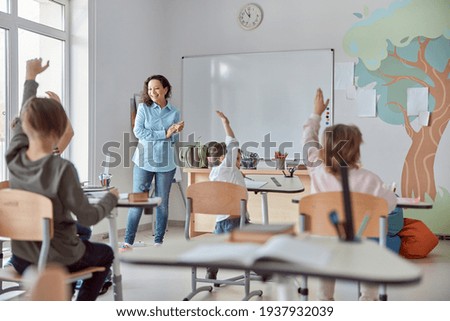 Young happy elementary school pupils at light modern classroom are rising their hands on a lesson Royalty-Free Stock Photo #1937932039