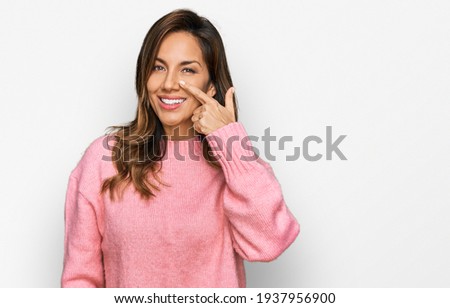 Young hispanic woman wearing casual clothes pointing with hand finger to face and nose, smiling cheerful. beauty concept  Royalty-Free Stock Photo #1937956900