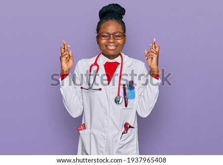 Young african american woman wearing doctor uniform and stethoscope gesturing finger crossed smiling with hope and eyes closed. luck and superstitious concept. 